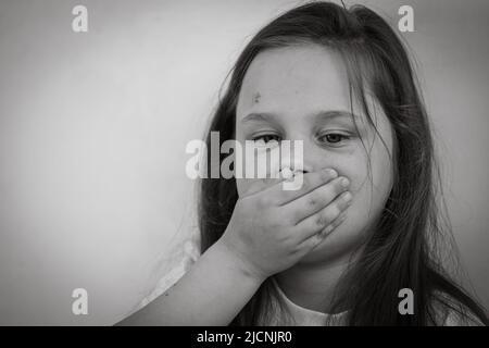 Ritratto bianco e nero di piccola ragazza triste livida con capelli lunghi e scuri che ricoprono la bocca con la mano. Protesta contro gli abusi. Foto Stock