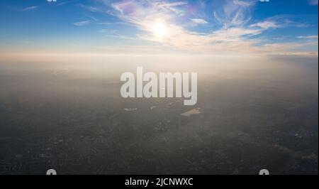 Vista aerea dalla finestra dell'aeroplano ad alta quota della città lontana coperta con strato di smog sottile e nubi distanti in serata Foto Stock