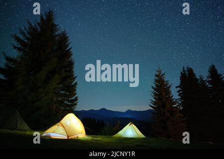 Luminose tende turistiche illuminate che brillano sul campeggio in montagne scure sotto il cielo notturno con stelle scintillanti. Concetto di stile di vita attivo Foto Stock