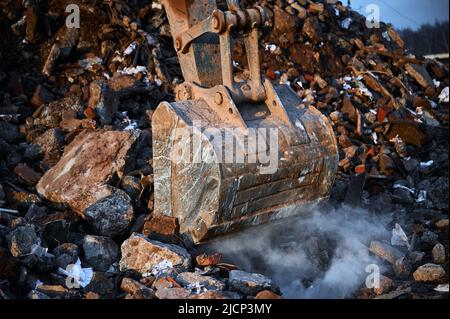 La benna di un escavatore da costruzione raccoglie calcestruzzo distrutto e mattoni e grandi pali. Foto Stock