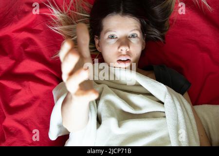 La ragazza si trova in un letto su un foglio rosso e allunga le braccia fino alla cima, dormire e riposare a casa a letto, comfort e intimità Foto Stock