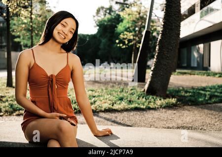 Giovane donna asiatica sorridente alla macchina fotografica mentre si rilassa seduto su una panca all'aperto. Foto Stock
