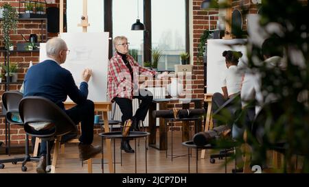 Artista anziano che spiega le abilità di disegno alla gente al programma della scuola d'arte, insegnandoli a disegnare lo schizzo professionale per la crescita personale. Laboratorio artistico di pratica hobby. Scatto su cavalletto Foto Stock