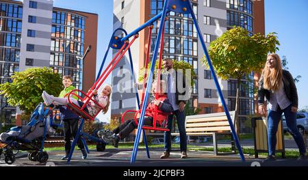 Famiglia felice - padre, madre e bambini che si divertono insieme sul parco giochi. I genitori oscillano figlia e figlio, mentre l'altro bambino in piedi con il pram. Moderni edifici residenziali sullo sfondo. Foto Stock