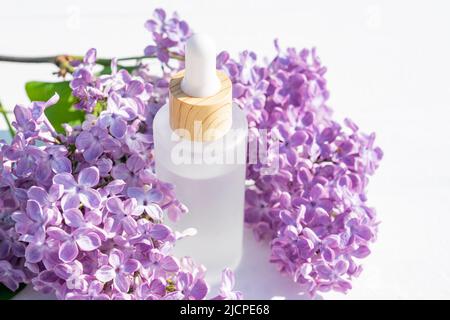 Essenza di siero o collagene liquido in bottiglia di vetro smerigliato su sfondo bianco con fiori viola lilla. Concetto di prodotti cosmetici anti age Foto Stock