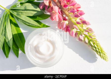 Crema cosmetica bianca senza marchio in un vaso aperto e fiori lupini luminosi su sfondo bianco. Mockup, modello. Contenitore per lozione corpo, idratante cr Foto Stock