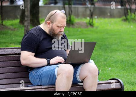 Uomo bearded in jeans pantaloncini seduti con un computer portatile su una panca nel parco estivo. Concetto di freelance che lavora all'aperto Foto Stock