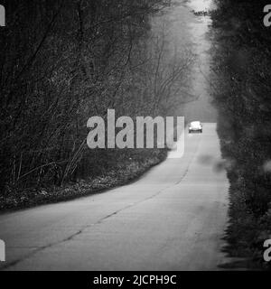 Auto con fari illuminati fuori fuoco che si avvicina lungo tortuoso asfalto strada di campagna circondato da fitta foresta autunno e nebbia nera e. Foto Stock