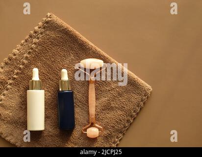 Set per la cura del viso a casa su un asciugamano marrone. Rullo per massaggio viso al quarzo rosa e bottiglie in vetro bianco e blu - siero notturno e diurno per la cura del viso. Foto Stock