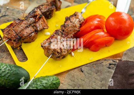 Carne di manzo alla griglia con spiedini d'acciaio su un tagliere con pomodori e cetrioli tagliati Foto Stock