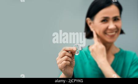 Donna matura che tiene l'apparecchio acustico BTE in mano in primo piano e punta all'orecchio. Trattamento della sordità in persone adulte Foto Stock