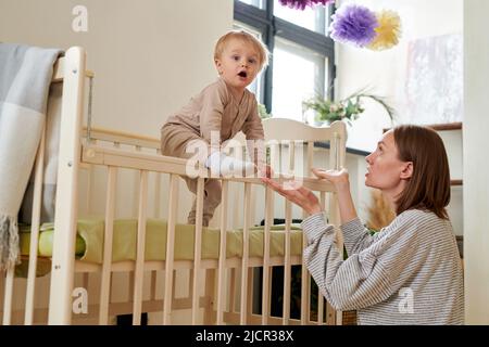 Il bambino si svegliò la mattina e si arrampicò fuori dal suo presepe con la madre che gli raggiunge le mani Foto Stock