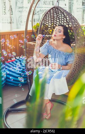 Una giovane donna con trucco luminoso è seduta in un caffè all'aperto estate in una sedia appesa e bolle di sapone soffiato. Foto Stock