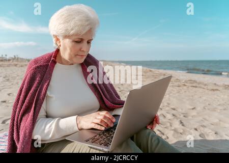 Donna anziana focalizzata che lavora sul computer in riva al mare Foto Stock