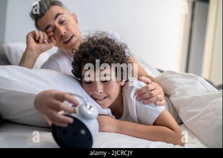 Un ragazzo carino dai capelli scuri che guarda l'orologio mentre rimane a letto Foto Stock