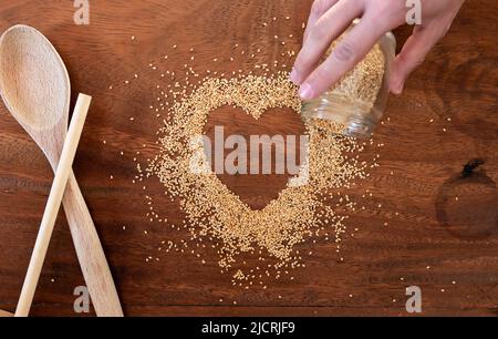 Semi di sesamo tostati biologici a forma di cuore su un piano di cottura in legno con utensili da cucina e una mano Foto Stock