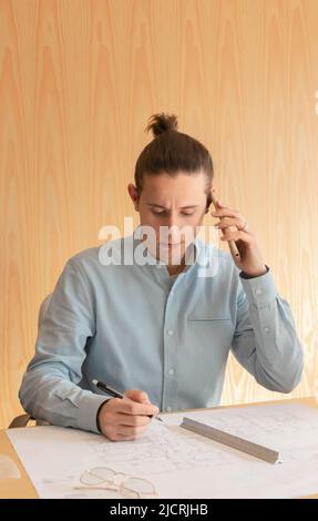 Uomo d'affari con espressione arrabbiata mentre parla al telefono e ha alcuni progetti architettonici sulla scrivania in ufficio Foto Stock