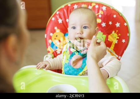 la madre nutre il bambino divertente da un cucchiaio. il bambino mangia in un seggiolone Foto Stock