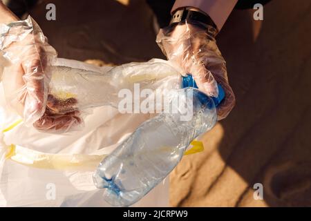 Mani umane tagliate in guanti di protezione che puliscono l'area sabbiosa da rifiuti di plastica. Raccogliere il flacone nel sacchetto. Problema ECO Foto Stock