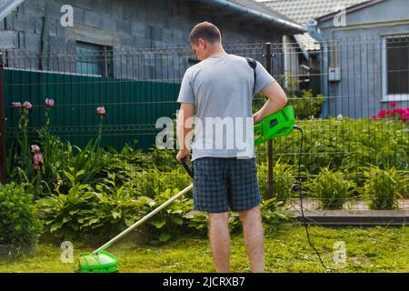 Rifinitore per erba. Vista posteriore di un uomo che rasa l'erba. Vista all'aperto di un giovane lavoratore che utilizza un rasaerba tagliando l'erba in uno sfondo sfocato natura Foto Stock