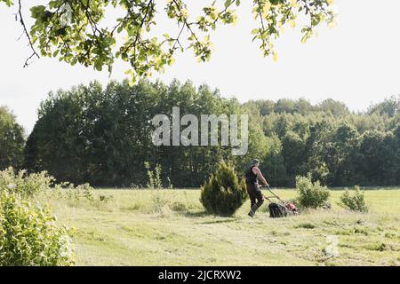 L'uomo rasa l'erba con un tosaerba elettrico nel giardino. Il proprietario che lavora duramente si occupa del prato in una giornata estiva soleggiata. Foto Stock