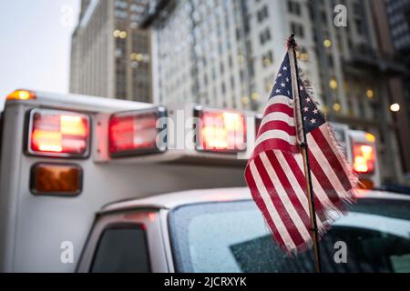 Primo piano della bandiera americana contro la luce lampeggiante rossa della sirena dell'auto dell'ambulanza. Temi di emergenza, soccorso e aiuto. Foto Stock