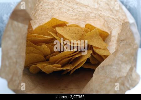 Sacchetto di carta marrone pieno di nachos gialli su un tavolo bianco. Antipasto in stile messicano. Cibo malsano ma delizioso per spuntini o mangiare. Croccante! Foto Stock