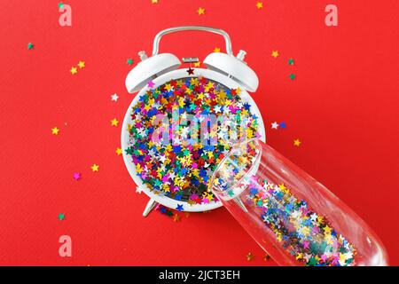 Un bicchiere con paillettes multicolore, sveglia e confettie a forma di stella sullo sfondo rosso. Festa, anno nuovo, concetto di festa Foto Stock