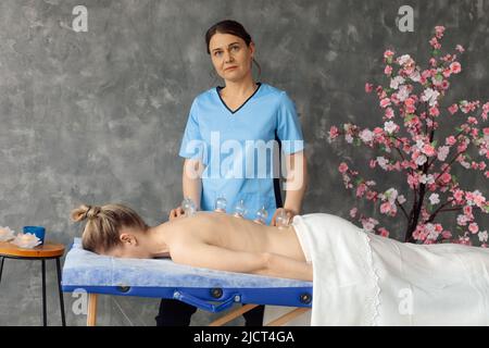 Massaggiatore in cabinet massaggiando i muscoli della schiena di una donna sdraiata sul divano con coppette da vuoto mediche. Ricevimento specializzato, massaggio professionale e. Foto Stock