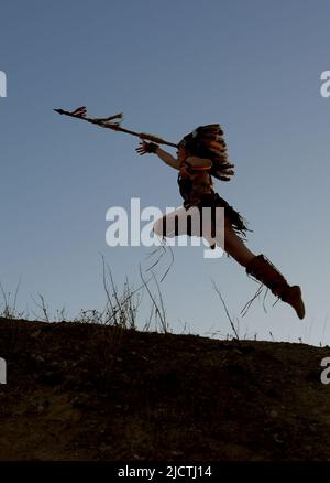 Una giovane ragazza è silhouetted come indiana americana natale. Si vede correre e caricare una collina al tramonto con una lancia in mano. Foto Stock