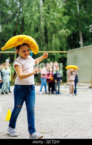 Sorridente bambina che porta su cuscino giallo nel parco giochi nel parco verde. Vacanze estive in campeggio, centro turistico. Passeggiate e giochi all'aperto Foto Stock