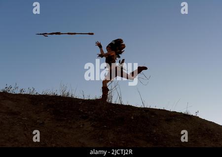 Una giovane ragazza è silhouetted come indiana americana natale. Si vede correre e caricare una collina al tramonto con una lancia in mano. Foto Stock