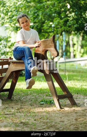 Carina bambina seduta su cavallo di legno nel parco giochi verde. Vacanze estive in campeggio, centro turistico. Passeggiate e giochi all'aperto, sport Foto Stock