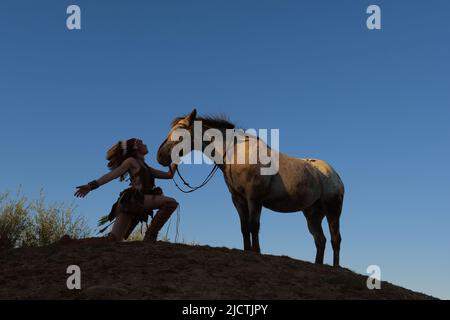 Una giovane ragazza è vista al tramonto come un indiano nativo americano. Si ferma sulla cima di una piccola collina per custodire il suo cavallo Foto Stock