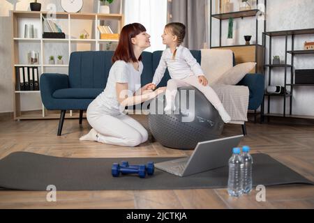 Media età bello sport caucasico con la sua figlia piccola carina che fa l'esercizio di yoga usando la palla fitness a casa. Giovane famiglia sportiva sorridendo felicemente allenando pilates utilizzando il fit. Foto Stock