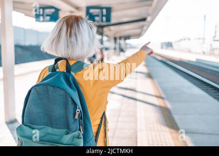 European Travel 50s femmina con viaggio zaino mostra la direzione per il turista via alla stazione della piattaforma ferroviaria Foto Stock