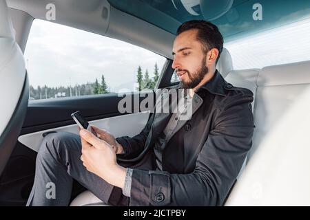 Giovane caucasico uomo di successo che parla seriamente sullo smartphone mentre si siede sul sedile posteriore di un'auto di guida Foto Stock