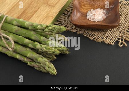 Mazzo di asparagi verdi legato con filo di iuta vicino al tagliere su sfondo nero con spazio copia. Cottura degli asparagi e concep di verdure primaverili Foto Stock