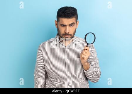 Ritratto di un uomo d'affari serio e sicuro che tiene in mano la lente d'ingrandimento e guarda la macchina fotografica, lo spionaggio, indossando la camicia a righe. Studio interno girato isolato su sfondo blu. Foto Stock