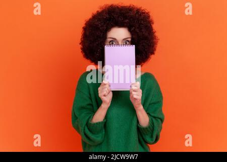 Ritratto di donna con acconciatura Afro indossando un maglione verde casual che copre metà del suo volto con carta organizer, guardando la macchina fotografica. Studio interno girato isolato su sfondo arancione. Foto Stock