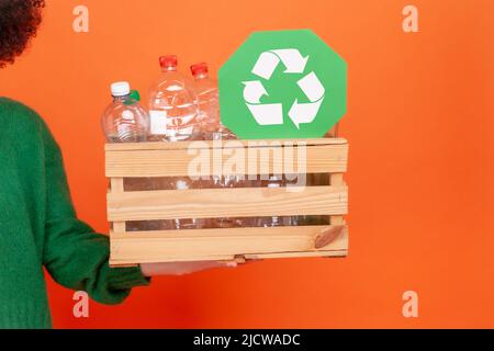 Mano di donna che indossa il maglione verde casual stile che tiene la scatola con bottiglie vuote di plastica e il simbolo verde di riciclaggio, problema di ecologia. Studio interno girato isolato su sfondo arancione. Foto Stock