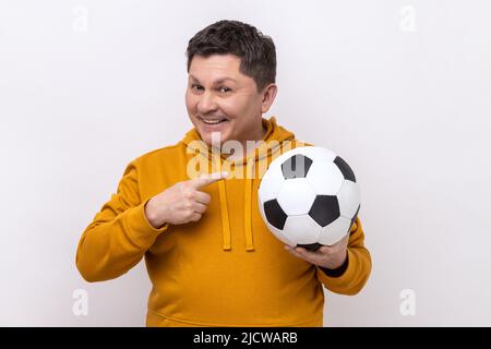 Bell'uomo di mezza età che punta il dito sulla palla di calcio in mano con espressione positiva sorridente, tifoso di calcio, indossando felpa con cappuccio in stile urbano. Studio interno girato isolato su sfondo bianco. Foto Stock