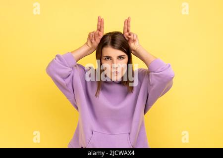 Ritratto di donna aggressiva che tiene le dita sopra la testa mostrando corna, arrogante e testardo, pronto ad attaccare, indossando felpa con cappuccio viola. Studio interno girato isolato su sfondo giallo. Foto Stock