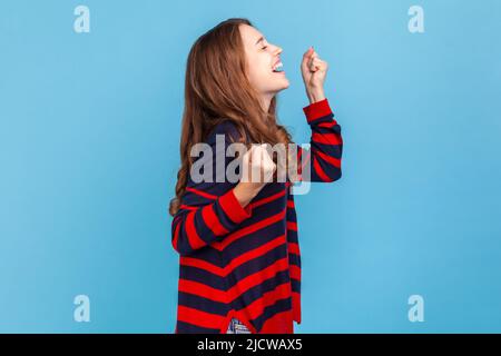 Vista laterale della donna felice indossando un pullover casual a righe in piedi con pugni clenched rialzati, celebrando la vittoria, esprimendo la vittoria. Studio interno girato isolato su sfondo blu. Foto Stock