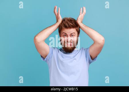 Ritratto di uomo positivo barbuto in piedi con corna di corna sopra la testa e mostrando lingua fuori, guardando con comica espressione umoristica. Studio interno girato isolato su sfondo blu. Foto Stock