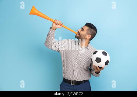 Un uomo d'affari felice celebra la vittoria della squadra di calcio preferita, soffiando il corno e tenendo la palla in mano, indossando una camicia a righe. Studio interno girato isolato su sfondo blu. Foto Stock