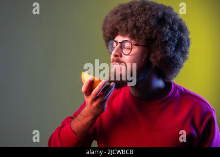 Hipster uomo con acconciatura Afro odorante mezza fetta di pompelmo, gustando gustosi frutti freschi succosi vitaminici, sana alimentazione dietetica. Studio interno girato isolato su colorato sfondo di luce al neon. Foto Stock