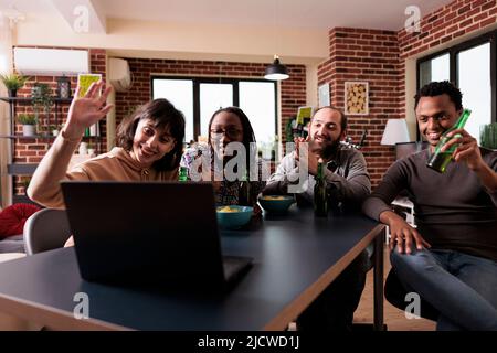 Gruppo multietnico di amici che sventolano la mano alla videochiamata online sul computer portatile. Diverse persone sedute a casa in soggiorno mentre fanno gesto di saluto alla conversazione su Internet. Foto Stock