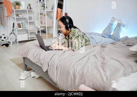Vista laterale ritratto di ragazza asiatica teen usando il laptop mentre si trova sul letto in stanza accogliente, spazio copia Foto Stock
