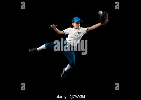 Ritratto dinamico di piccolo giocatore di baseball, caraffa in uniforme blu-bianco formazione isolato su sfondo nero studio. Concetto di sport, realizzazioni Foto Stock